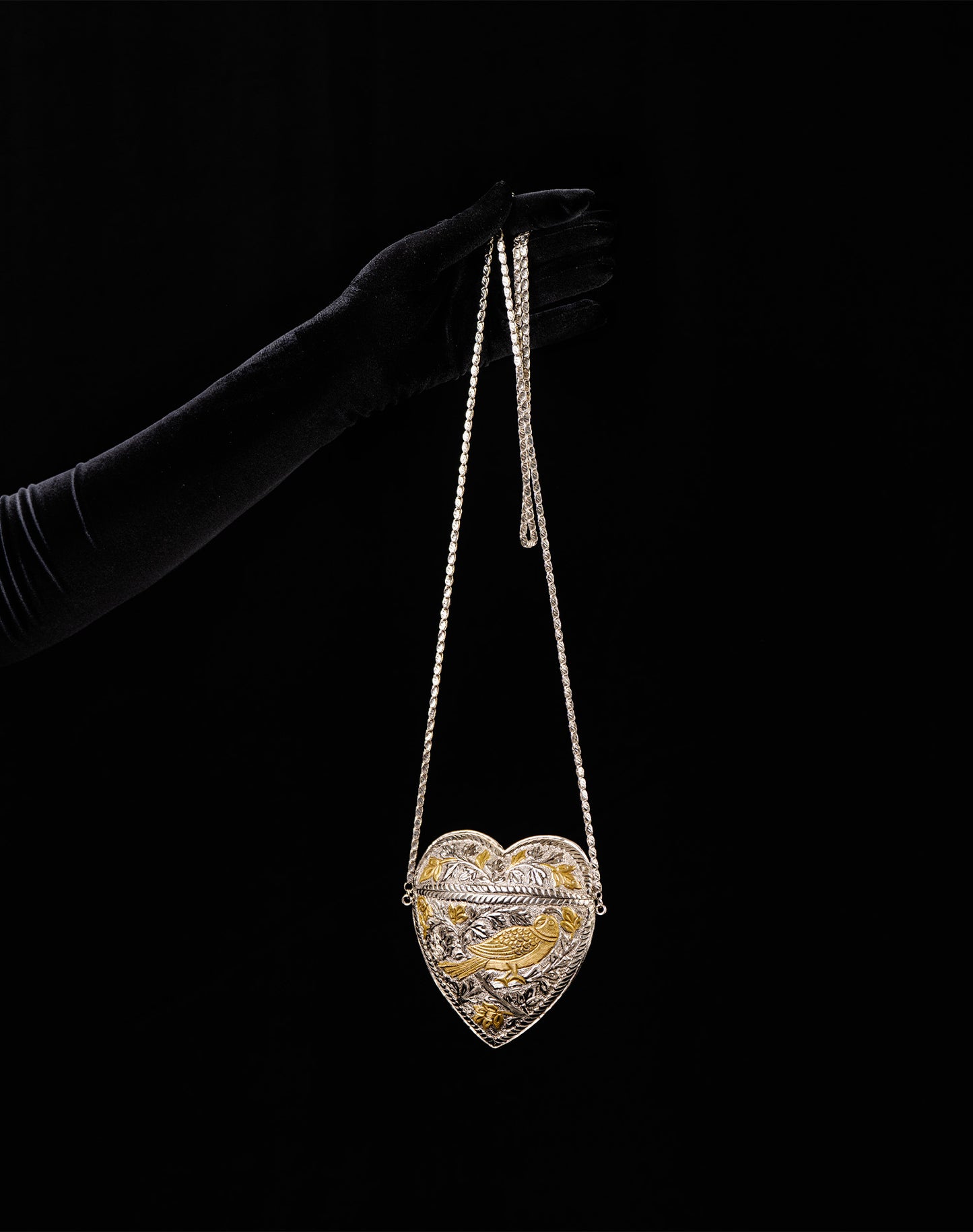 Heart-shaped jewelry box with gold accents held by a gloved hand against a black background